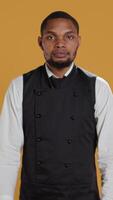 Vertical Waiter flexing his muscles and showing his strength in studio, acting tough while he wears his restaurant uniform and apron. Professional employee in service oriented industry. Camera B. video