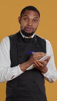 Vertical Skilled waiter with apron takes meal orders from clients on a notebook, writing down important information for customer satisfaction. Caterer butler working in service oriented industry. Camera B. video