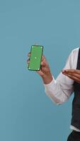 Vertical Hotel worker bellhop points at phone with isolated mockup display, showing green screen against blue background in studio. Hotelier doorman greeting resort guests at reception. Camera B. video