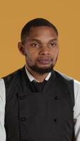 Vertical Waiter flexing his muscles and showing his strength in studio, acting tough while he wears his restaurant uniform and apron. Professional employee in service oriented industry. Camera A. video