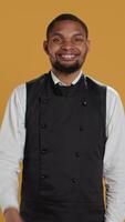 Vertical Waiter smiling and showing a thumbs up symbol against yellow background, presenting his satisfaction and approval with a positive like sign. Cheerful staff gives okay gesture. Camera B. video