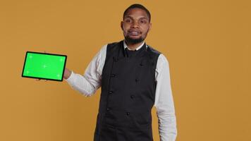 Catering employee presenting an isolated chroma key display while he wears an apron and serving clients at diner. Hospitality industry staff in professional attire, customer satisfaction. Camera B. video