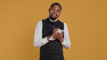 Professional caterer writing clients orders on notepad for the bill, takes notes of desired food against yellow background. Waiter with apron, notebook service for customer satisfaction. Camera B. video