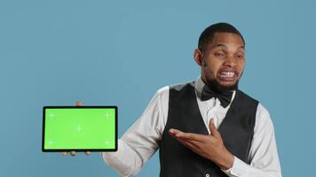 Doorkeeper bellboy poses with green screen on tablet at the hotel reception, working in hospitality industry and greeting customers. Lodging resort personnel shows chroma key. Camera A. video