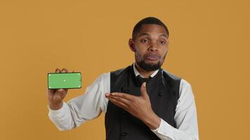 Waiter with professionalism holding a phone with isolated mockup display, working in dining and eatery industry. Friendly restaurant staff does customer oriented activity at his job. Camera A. video