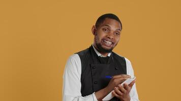 Professional caterer writing clients orders on notepad for the bill, takes notes of desired food against yellow background. Waiter with apron, notebook service for customer satisfaction. Camera A. video