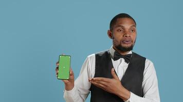Hotel worker bellhop points at phone with isolated mockup display, showing green screen against blue background in studio. Hotelier doorman greeting resort guests at reception. Camera A. video