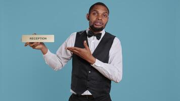 Hotel concierge staff showing the reception indicator at front desk, greeting guests at five star holiday resort in studio. Skilled professional bellboy holds sign against blue background. Camera B. video