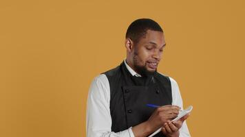 Skilled waiter with apron takes meal orders from clients on a notebook, writing down important information for customer satisfaction. Caterer butler working in service oriented industry. Camera A. video
