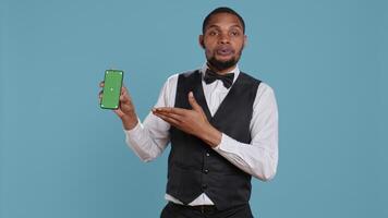 Hotel worker bellhop points at phone with isolated mockup display, showing green screen against blue background in studio. Hotelier doorman greeting resort guests at reception. Camera B. video