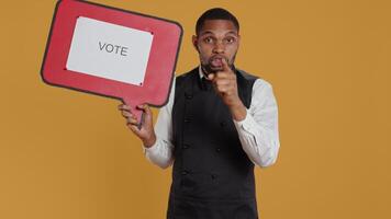 Catering employee showing a message to vote on red speech bubble, presenting the cardboard icon to do a commercial for politics. Waiter in uniform works on customer service. Camera B. video