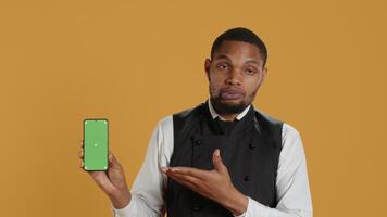 Restaurant staff showing a smartphone with green screen in studio, prepared to serve food orders in service oriented environment. Waiter with apron and uniform serving clients at tables. Camera A. video