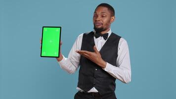 Bellman hotelier showing a tablet with green screen display in studio, presenting mockup chroma key screen against blue background. Young hotel concierge works at resort. Camera B. video