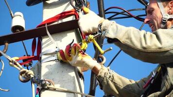 Skilled electrician in helmet fixes wires standing on ladder near high pole against blue sky on summer day backside view. Electrical service and mounting on the pole. Slow motion video