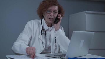 Middle-aged Caucasian female doctor sitting in her consulting room using phone and working on laptop. Elderly general practitioner talking on phone with patient while working on computer in clinic. video