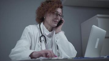 Cheerful mature woman doctor working with laptop, talking by phone in office. Connected with patient. Medicine, people and healthcare concept. General practitioner talking to colleagues by smartphone. video