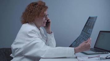 Elderly caucasian woman doctor analyze x-ray image and talk with mobile phone about diagnostics in medical office. Doctor specialist working with CT scan, talk to cellphone about radiography results. video