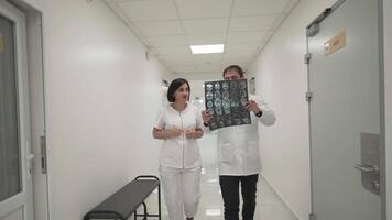 Teamwork in hospital. Two doctors move quickly across corridor of clinic and discuss an MRI scan of head of patient. Head doctor and assistant discuss CT scan of brain tumor patient in the hallway. video