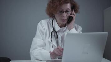 Middle-aged Caucasian female doctor sitting in her consulting room using phone and working on laptop. Elderly general practitioner talking on phone with patient while working on computer in clinic. video