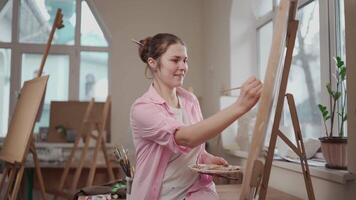 Cute woman paints on canvas in an art workshop. Artist creating picture. Art school or studio. Work with paints, brushes and easel. Hobby and leisure concept. Woman painter at workspace. video