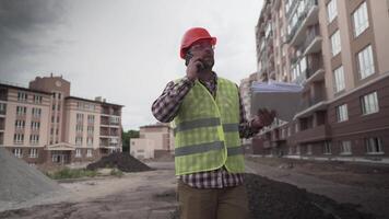 Builder in protective work clothing walks through construction of houses talking on phone with cbuilding plan of objects in hands. Foreman controls construction process while walking through project. video
