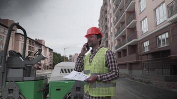 Engineer talking on phone with documents and plans in hands in front construction equipment at building site of apartment block. Supervisor in hard hat with heavy machinery at development site. video