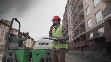 Foreman in hard hat, safety jacket talking by phone with building papers in hand in front construction machinery in apartment house complex site. Male architect outdoors at worksite with cell phone. video