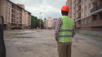Confident engineer building architect in hard hat stands outside with his back to camera at construction site. Man foreman in helmet and safety vest standing back and looking ahead at the worksite. video