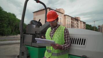 Civil engineer on construction site holding blueprints and smartphone in front of bulldozer for paving asphalt. Concept of apartment block constructions. Builder with roadmap near machinery. video