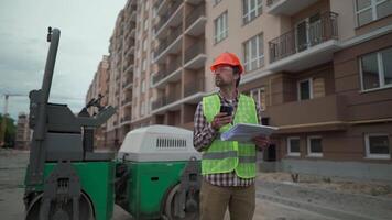 Engineer builder hard hat standing against paver at construction site, checking plan of project with technical documentation and using smartphone. Worker in front of roller on building site. video