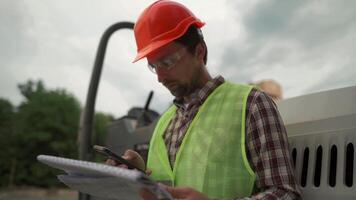Engineer builder hard hat standing against paver at construction site, checking plan of project with technical documentation and using smartphone. Worker in front of roller on building site. video