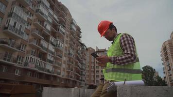 Builder relaxes sitting on construction elements at worksite, drinking coffee and surfing Internet on smartphone. Architect in helmet relaxing at work and drinking coffee too go with phone in hand. video
