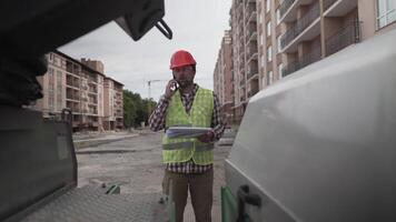 Engineer talking on phone with documents and plans in hands in front construction equipment at building site of apartment block. Supervisor in hard hat with heavy machinery at development site. video