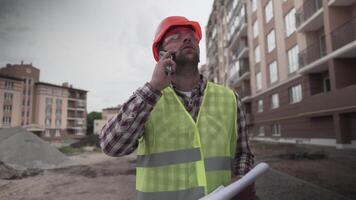 Working process. Contractor in working uniform control according to plan. Builder phone call and walking along construction site of new residential area. Foremant supervising construction. video