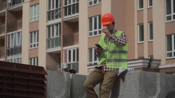 Builder relaxes sitting on construction elements at worksite, drinking coffee and surfing Internet on smartphone. Architect in helmet relaxing at work and drinking coffee too go with phone in hand. video