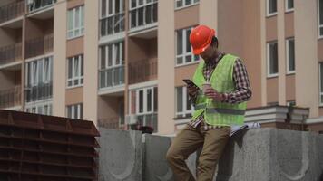 Construction worker resting on building site drinking coffee to go and using smartphone while sitting on jobsite. Architect chatting and using smartphone during coffee break at work project location. video