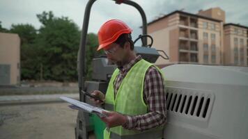 Inspection of civil construction at build site of high rise apartment house. Engineer examines documentation, plan and building progress on drawings in front of machine. Worker against of a paver. video
