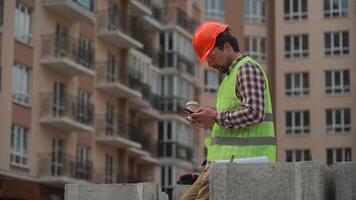 Construction industry worker texting during lunch break. Helmeted handyman chatting and using smartphone during coffee break at worksite. Architect in hard hat and vest drinks coffee at building site. video