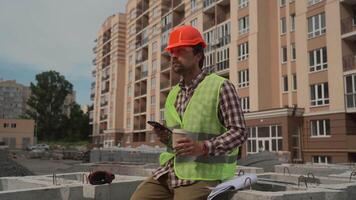 Construction worker resting on building site drinking coffee to go and using smartphone while sitting on jobsite. Architect chatting and using smartphone during coffee break at work project location. video
