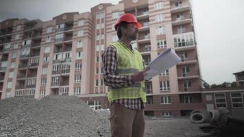 Construction inspector with blueprints on building site. Engineer in protective workwear checking technical design project construction of apartment buildings. Architect examining building plans. video
