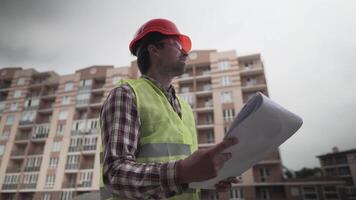 Construction inspector with blueprints on building site. Engineer in protective workwear checking technical design project construction of apartment buildings. Architect examining building plans. video