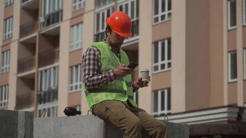 Builder relaxes sitting on construction elements at worksite, drinking coffee and surfing Internet on smartphone. Architect in helmet relaxing at work and drinking coffee too go with phone in hand. video