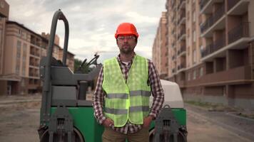 Portrait construction worker in protective overalls proudly posing against asphalt paver at construction site. Builder in hard hat and vest looks at camera and smile background of asphalt road roller. video