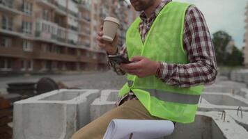 Construction worker resting on building site drinking coffee to go and using smartphone while sitting on jobsite. Architect chatting and using smartphone during coffee break at work project location. video