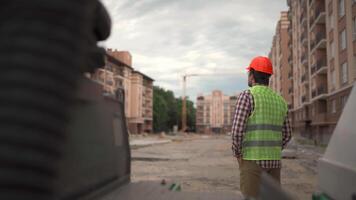Back view of male construction worker in orange hard hat and uniform looking at building site of an apartment building at the end of the work day. Theme architecture and civil engineering. video