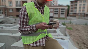 Builder relaxes sitting on construction elements at worksite, drinking coffee and surfing Internet on smartphone. Architect in helmet relaxing at work and drinking coffee too go with phone in hand. video
