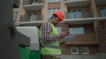 Civil engineer on construction site holding blueprints and smartphone in front of bulldozer for paving asphalt. Concept of apartment block constructions. Builder with roadmap near machinery. video