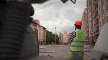 Theme architecture and civil engineering. Young male inspector at construction site standing back to camera and inspecting building area of housing estate without workers at end of the work day. video