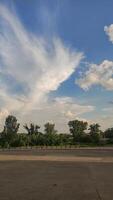 A blue sky with fluffy white clouds above a row of green trees and an empty parking lot photo