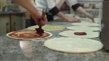 A group of cooks evenly put tomato sauce on pizza dough circles. Close-up video